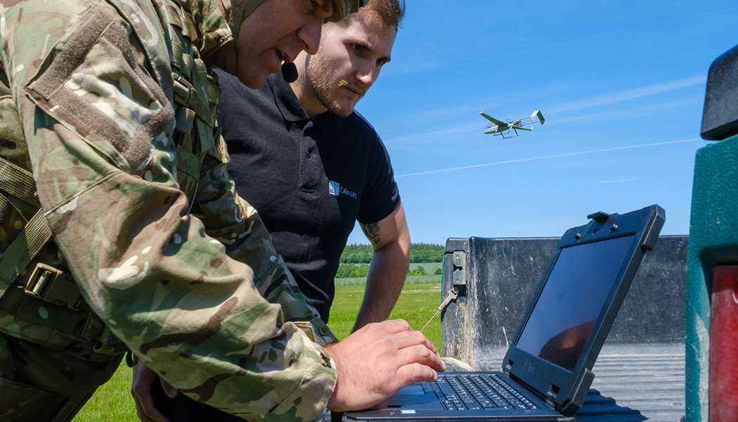Military personnel using a laptop and a drone flying in the background