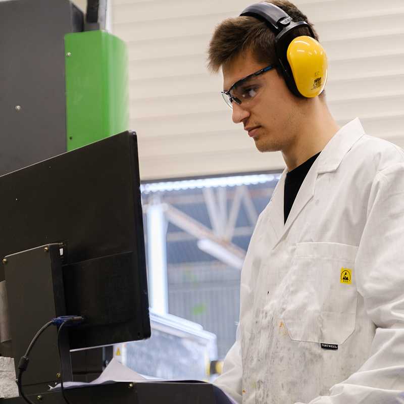 Engineer in white coat and yellow ear defenders observing a monitor